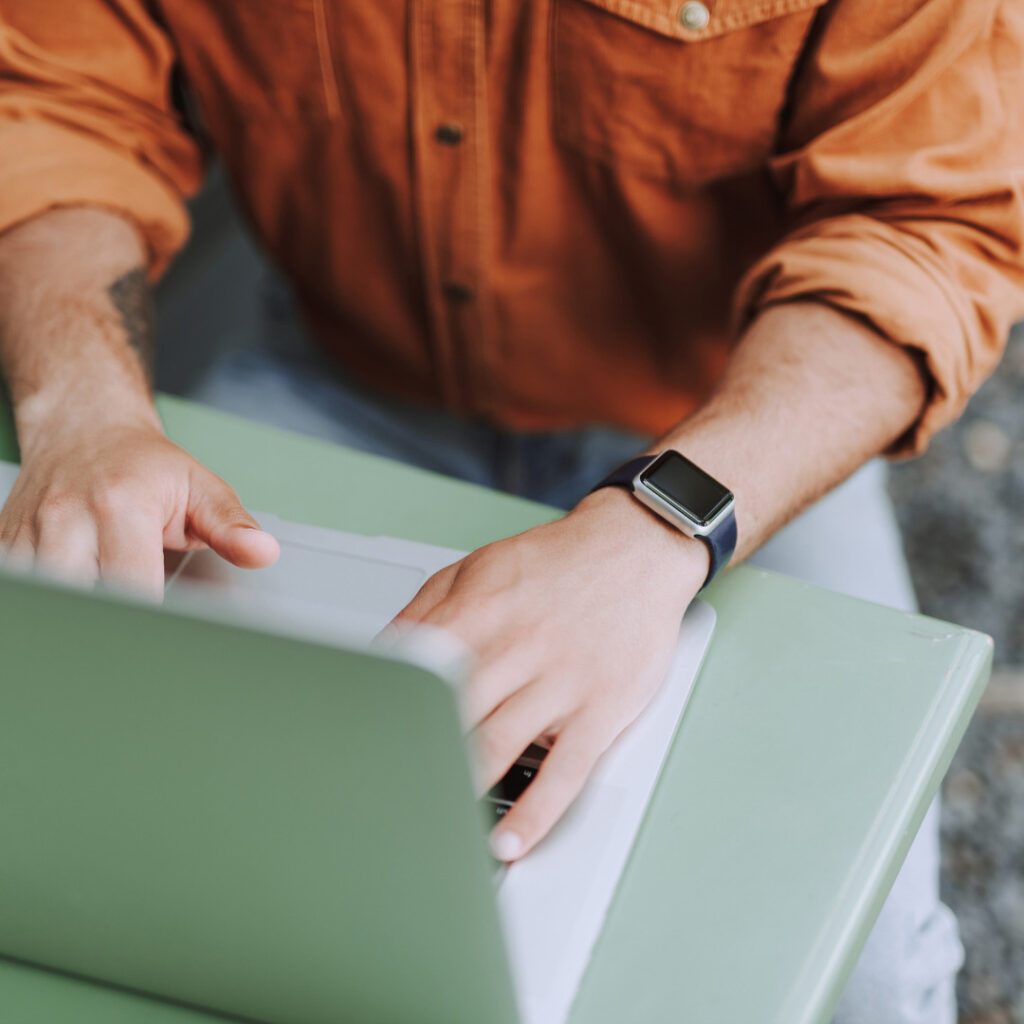 
Mies istuu vihreän pöydän ääressä ja kirjoittaa kannettavalla tietokoneellaan. Hänellä on yllään oranssi paita ja vasemmassa ranteessaan älykello. Kuvassa näkyvät lähinnä hänen kädet ja osa ylävartaloa.