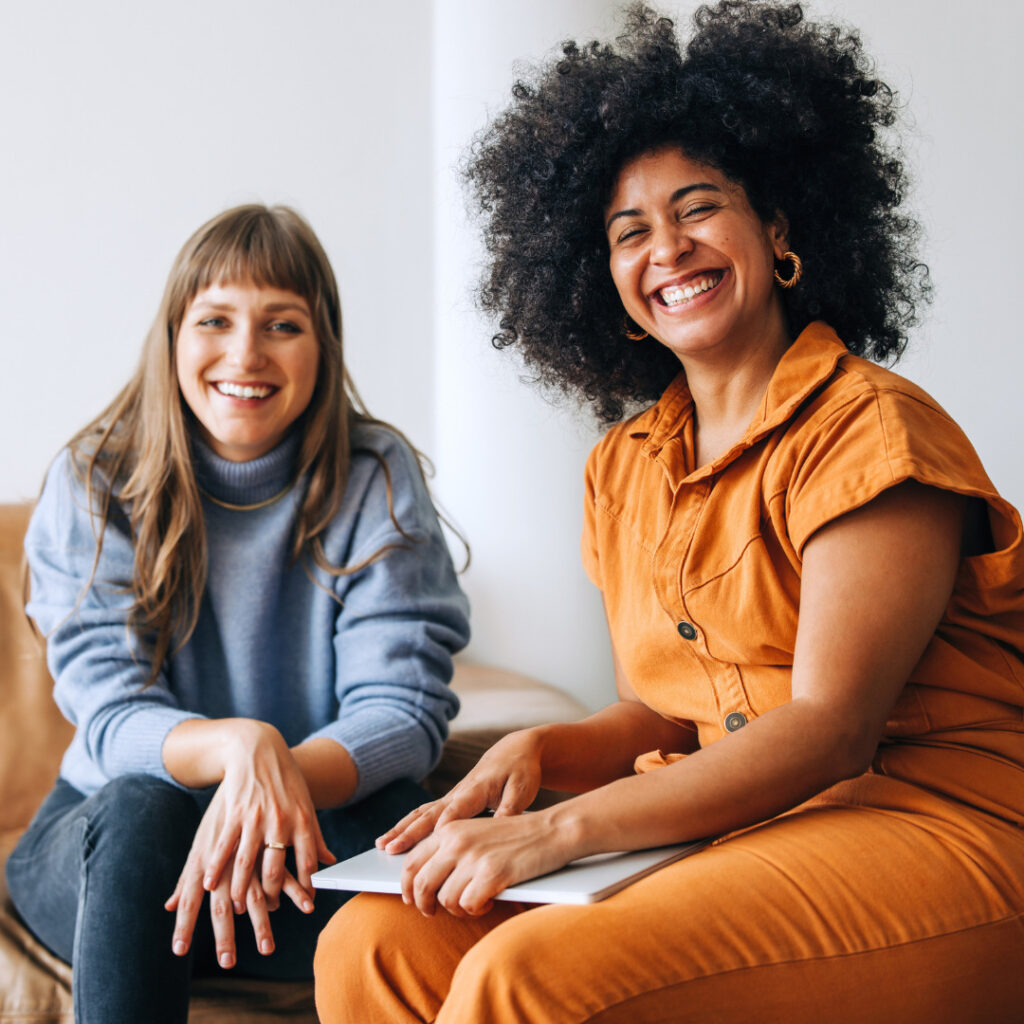 Kaksi naista istuu sohvalla ja katsoo kameraan, leveästi hymyillen. Vasemmalla oleva nainen on vaaleahiuksinen ja hänellä on yllään vaaleansininen neule. Oikealla oleva naisella on suuret afrohiukset ja oranssi haalari. Hänellä on sylissään kannettava tietokone.