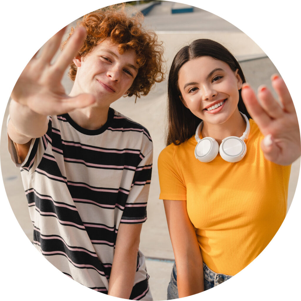 
Pyöreään muotoon rajattu kuva kahdesta nuoresta, jotka seisovat ulkona ja katsovat kameraan. Vasemmalla oleva nuori, jolla on punertavat, kiharat hiukset ja raidallinen paita, nostaa toisen kätensä kohti kameraa. Oikealla oleva nuori, jolla on pitkät tummat hiukset ja keltainen T-paita, hymyilee leveästi ja pitää valkoisia kuulokkeita kaulallaan, myös hän nostaa kätensä kohti kameraa. Molemmat vaikuttavat iloisilta.